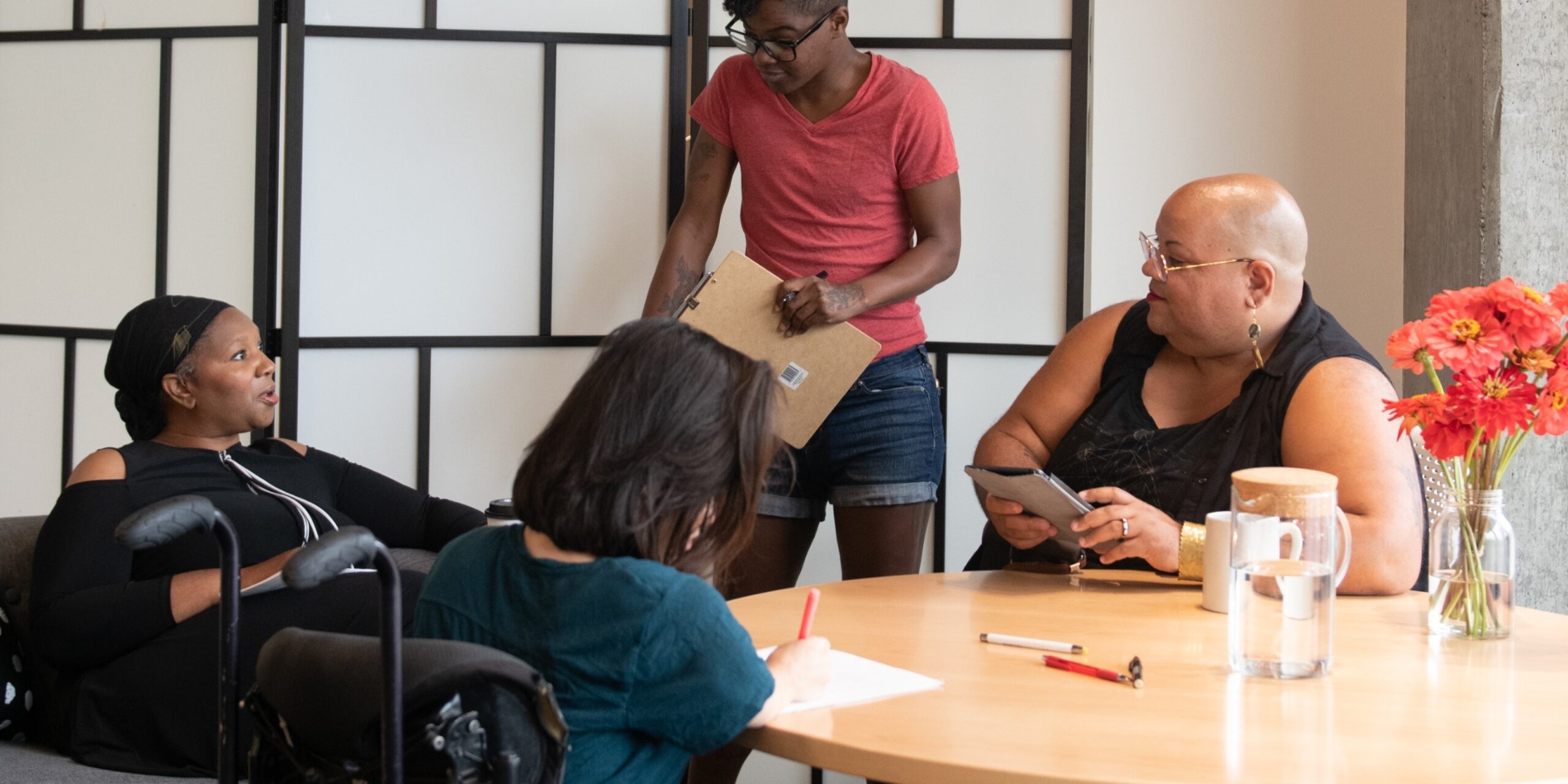 Vier mensen van kleur met een beperking zitten samen aan tafel tijdens een vergadering. Een zwarte vrouw zit op een bank en spreekt met een neutrale gezichtsuitdrukking. De drie anderen luisteren: een Zuid-Aziatisch persoon in een rolstoel maakt aantekeningen, een zwarte non-binaire persoon zit in een stoel met een tablet en een wandelstok, en een andere zwarte non-binaire persoon staat met een clipboard in de hand. Als illustratie bij het artikel over validisme.