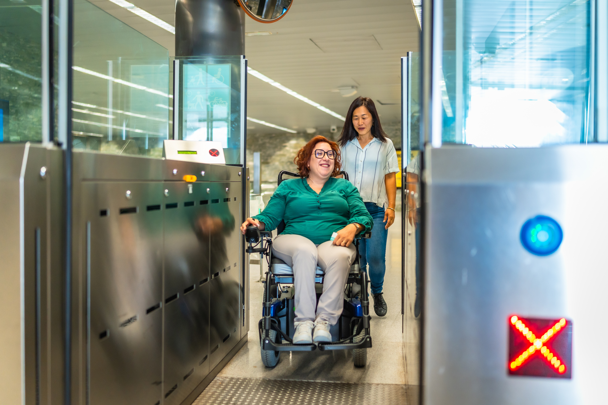 Een lachende witte vrouw in een rolstoel en een Oost-Aziatische begeleider gaan samen door een metropoortje als voorbeeld van universeel ontwerp